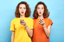 Surprised Emotional Twins With Wide Open Mouth And Bugged Eyes Holding Smart Phones And Looking At The Camera. Sale, Business, Free Message, Double Discounts