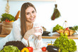 © rogerphoto - Young happy woman is holding white cup and looking at the camera while sitting at wooden table in the kitchen among green vegetables