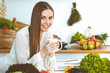 © rogerphoto - Young happy woman is holding white cup and looking at the camera while sitting at wooden table in the kitchen among green vegetables