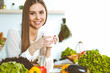 © rogerphoto - Young happy woman is holding white cup and looking at the camera while sitting at wooden table in the kitchen among green vegetables
