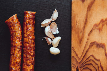 Two sausages and garlic next to empty wooden board – Pork meat and cloves on black ardesia stone