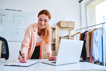 Young woman marking deliveries on clipboard and looking at laptop in office