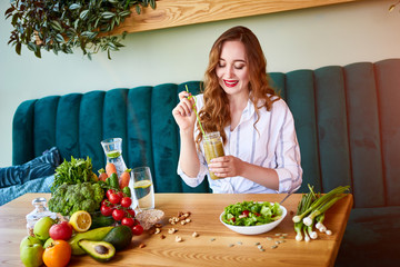 Wall Mural - Young woman drinking smoothie in the beautiful interior with green flowers on the background and fresh fruits and vegetables. Healthy eating concept. Vegan meal and detox menu