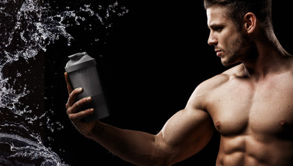 Wall Mural - Model sports young man on dark background. Portrait of beautiful strong muscle guy with gray protein drink in shaker. Water splash.