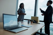 © DragonImages - Opened laptop and mug of coffee on office table, talking business people in background