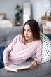 © BestForYou - Young woman on sofa at home reading a book.