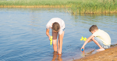 a girl and a boy take water from a lake into water guns in order to play them, against the backdrop of the landscape