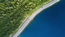Beautiful Aerial View Of Road Between Green Summer Forest And Blue Lake In Lapland. Car Moving On Road.
