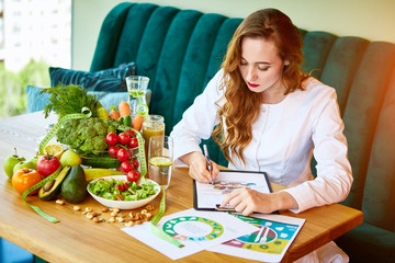 Woman dietitian in medical uniform with tape measure working on a diet plan sitting with different healthy food ingredients in the green office on background. Weight loss and right nutrition concept