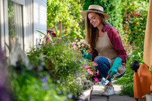 Portrait Of Happy Gardening Woman In Gloves, Hat And Apron Plants Flowers On The Flower Bed In Home Garden. Gardening And Floriculture. Flower Care