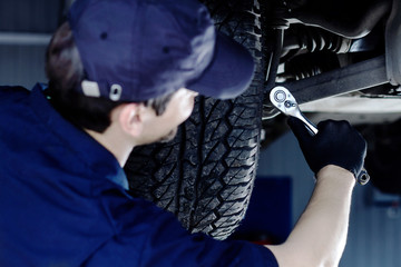 Mechanic in blue jumpsuit is repairing car at service station garage. Back view repairman is tightening nut by steel wrench at workshop auto repair shop. Vehicle on hydraulic lift is above.