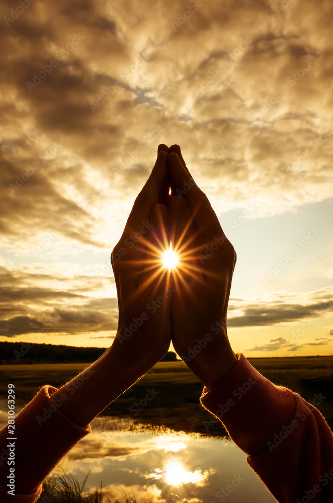 Tin namaste close-up, the sun shines between the fingers with stunning rays. Silhouette against the setting sun, sky and river. Concept of amateur yoga, healthy lifestyle, meditation practice.  - obrazy, fototapety, plakaty 