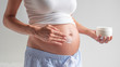 Authentic close up shot of an young pregnant woman in pajamas is applying a moisturizer or sun protection cream on her belly isolated on a white background.