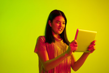 Wall Mural - Beautiful female half-length portrait isolated on yellow studio background in neon light. Young emotional woman. Human emotions, facial expression concept. Using tablet for talking or internet serfing
