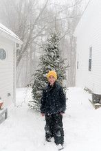 Smiling Little Boy With Yellow Hat Standing In Front Of Christmas Tree On Snowy Day