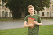 © bodnarphoto - Young smiling male student standing on the lawn in college park holding take away coffee and notebooks. Successful cheerful university student with a broad smile near the campus with coffee and books.