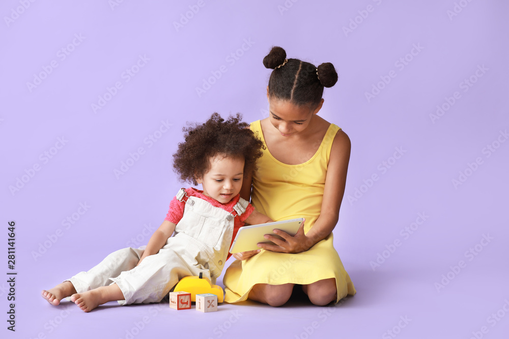 Cute African-American sisters with tablet computer watching cartoons on color background