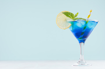 Bright summer fresh blue fruit cocktail with blue curacao liquor, ice cubes, lemon slice, green mint in pastel mint color interior on white wood board.