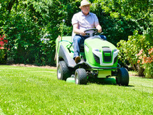 Senior Man Driving A Tractor Lawn Mower In Garden With Flowers