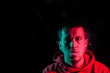 © Aleksandr Kondratov - Close up portrait of the face of an adult serious man exhales green toxic smoke while smoking e-cigarette and vape illuminated with blue and red colored light on a black background. Harm to health.
