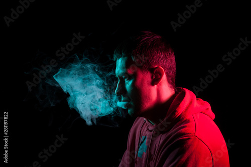 Cuadros en Lienzo Close up portrait of the face of an adult serious man exhales green toxic smoke while smoking e-cigarette and vape illuminated with blue and red colored light on a black background