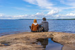 © Tatyana_Andreyeva - Tourists man and woman sitting on northern lake shore in summer day. People relaxing and admiring beautiful landscape. Travelling and discovering distant places of Earth. Onega lake, Karelia, Russia