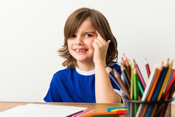 Little boy painting and doing homeworks on his desk showing a disappointment gesture with forefinger.