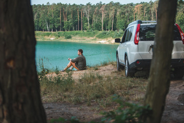 Wall Mural - man sitting near white suv car at the edge looking at lake with blue water