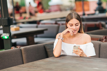 Lovely young woman holding a smartphone in her hands while sitting at a table in a street cafe