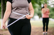 Motivation, weight loss, active lifestyle, group training, personal coach. Fat woman measuring her waistline with sporty female jogger working out at background