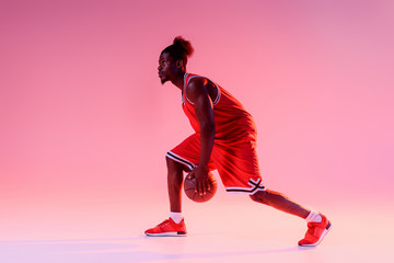 Wall Mural - handsome african american sportsman playing basketball on pink background with gradient and lighting