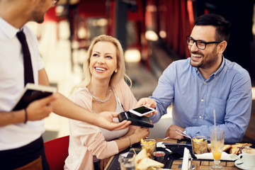 Happy couple paying a bill with smart phone on card machine in a restaurant.