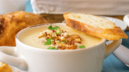 Blue wooden table with 2 bowls of cauliflower soup embellished with crushed hazelnuts, red pepper, smooth parsley herbs, Swiss twisted bread, frying pan with cauliflower, mortar and yellow flowers.