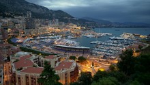 Dusk in Monaco. Cars are going the streets, boats and ships are moored by the shore of Mediterrenean Sea. Houses and streets are shining with orange city lights. The mountains are high above the city