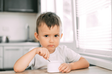 Wall Mural - Cute boy in the kitchen eating yogurt from a white yogurt container, a place for advertising