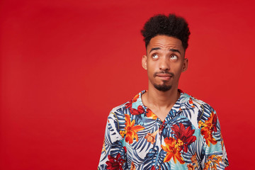 Wall Mural - Young pensive African American guy wears in Hawaiian shirt, looking up, thinking and imagine, stands over red background with copy space at the left side.