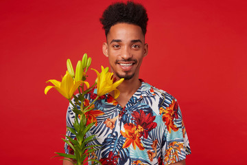 Wall Mural - Portrait of cheerful young African American guy, wears in Hawaiian shirt, looks at the camera with happy expression, stands over red background with yellow flowers and broadly smiles.