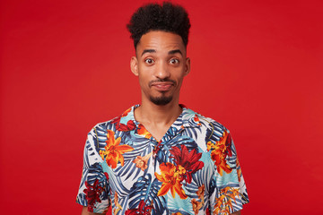 Wall Mural - Young confused African American guy wears in Hawaiian shirt, looking at the camera with wondered and misunderstanding, stands over red background with crossed arms.