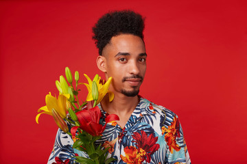 Wall Mural - Close up of calm young African American man, wears in Hawaiian shirt, looks at the camera, holds yellow and red flowers, stands over red background.