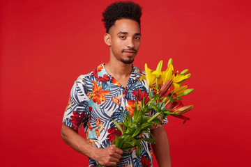 Wall Mural - Portrait of cheerful young African American man, wears in Hawaiian shirt, looks at the camera and smile, holds yellow and red flowers, stands over red background.