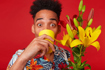 Wall Mural - Close up of young African American man in Hawaiian shirt, surprised looks at the camera and drinking water from a yellow glass, holds yellow and red flowers bouquet, stands over red background.