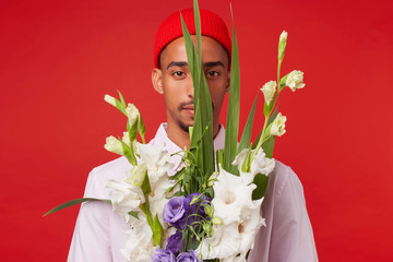 Wall Mural - Photo of young calm dark skinned man, wears in white shirt and red hat, looks at the camera trough bouquet , stands over red background.