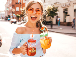 Young beautiful smiling hipster woman in sunglasses.Girl in summer trendy clothes posing in the street.Model holding and drinking two fresh cocktail smoozy drink