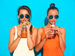 Two beautiful sexy smiling hipster women in summer colorful swimwear bathing suits.Trendy girls in sunglasses.Going crazy.Funny models isolated on blue.Drinking fresh cocktail smoozy drink