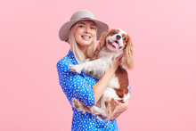 Cheerful Pretty Young Woman In Hat Hugging Her Dog On Pink Isolated Background. Lady Owner In Stylish Blue Dress And Braces. The Concept Of A Healthy Smile And Animal Care