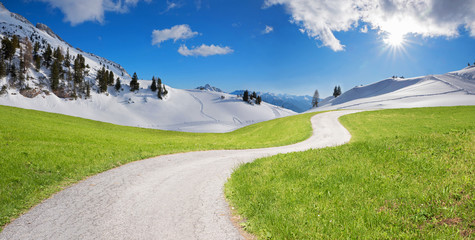 Fotomontage Klimawandel, grüne Wiese mit Kurvenweg und Winterlandschaft im Gebirge und Sonnenschein