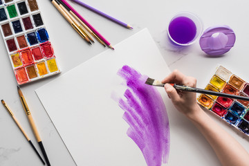 cropped view of artist drawing purple watercolor brushstrokes on white paper on marble white surface