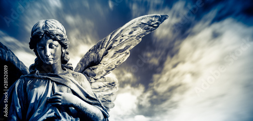 Guardian angel sculpture with open long wings isolated with blurred white clouds dramatic light blue sky. Angel sad expression sculpture with eyes down and hand in front of chest.