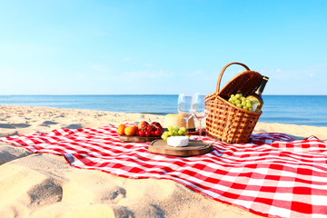 Checkered blanket with picnic basket and products on sunny beach. Space for text