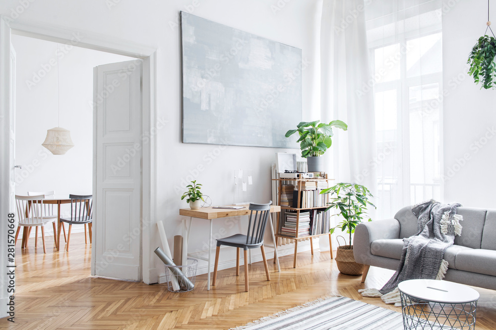 Stylish scandinavian open space with design furniture, plants, bamboo bookstand and wooden desk. Brown wooden parquet. Abstract painting. Modern decor of bright room next to dining room.  - obrazy, fototapety, plakaty 